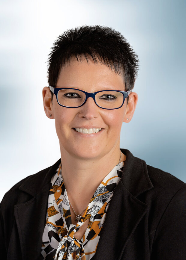 Simone Vogel, Geschäftsführung Sekretariat, hat kurze schwarze Haare. schwarze Brille, trägt eine bemusterte Bluse und ein schwarzes Jacket.