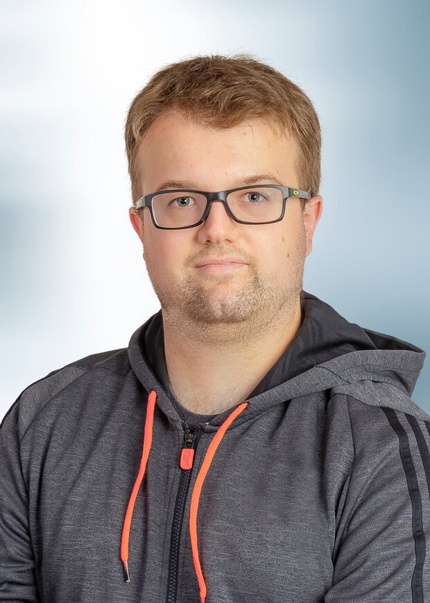 Dennis Kleinert, Verwaltung, hat blonde Haare, trägt eine Brille und einen grauen Hoodie.