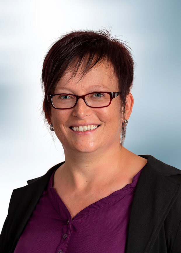 Jessica, Anger, Verwaltung, hat dunkelbraune Haare, trägt eine Brille und eine lila Bluse.