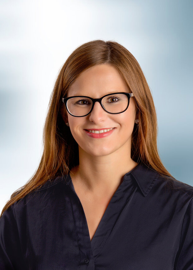 Alina Drochin, Disposition / Logistik, hat braune lange Haare, trägt eine Brille und eine dunkelblaue Bluse.