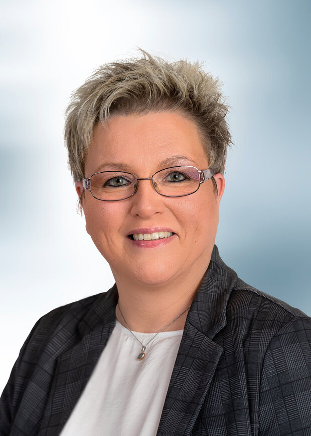 Petra Hilleke, Verwaltung, hat kurze blonde Haarte, trägt eine Brille mit einer weißen Bluse und einem grau-kariertem Blazer.