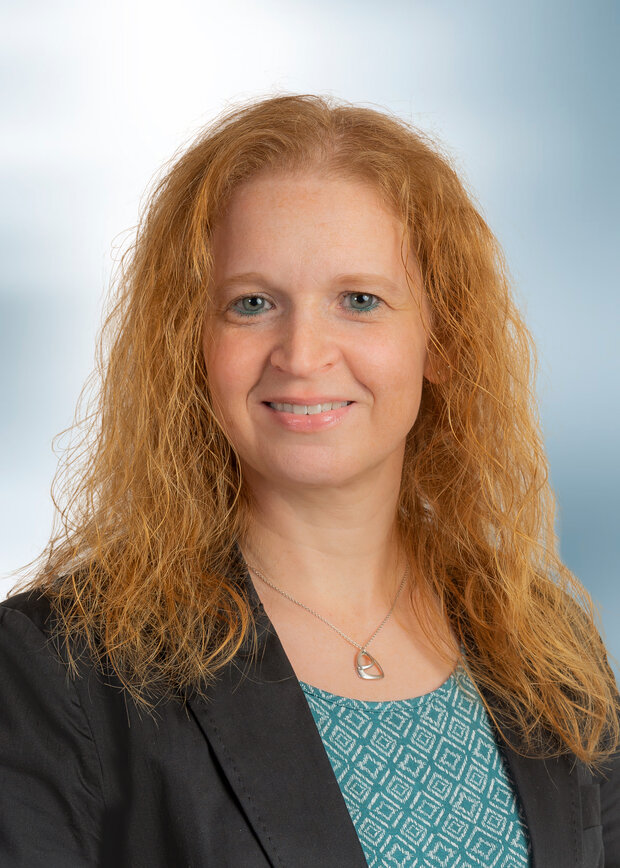 Janet Artus, Vertrieb Innendienst, hat rote, lange, lockige Haare und trägt ein blaues T-Shirt mit grauem Jacket.