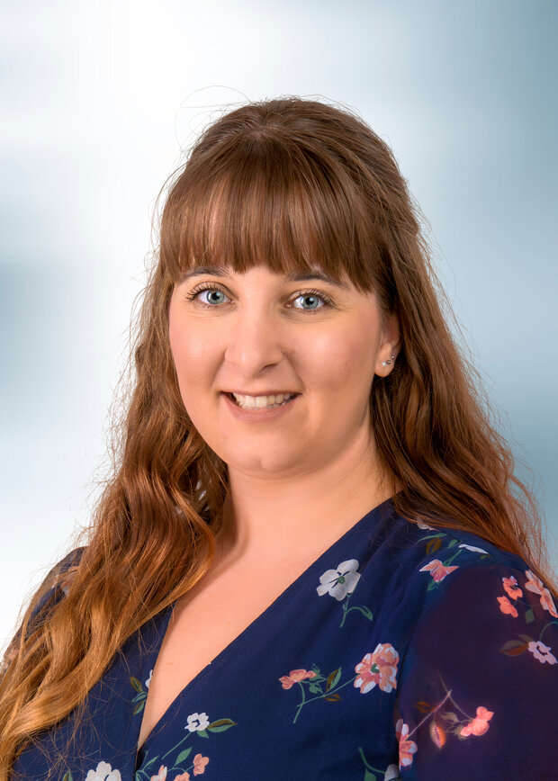 Jessica Hartmann, Marketing, hat lange braune Haare mit einem Pony und trägt eine dunkelblaue geblümte Bluse.